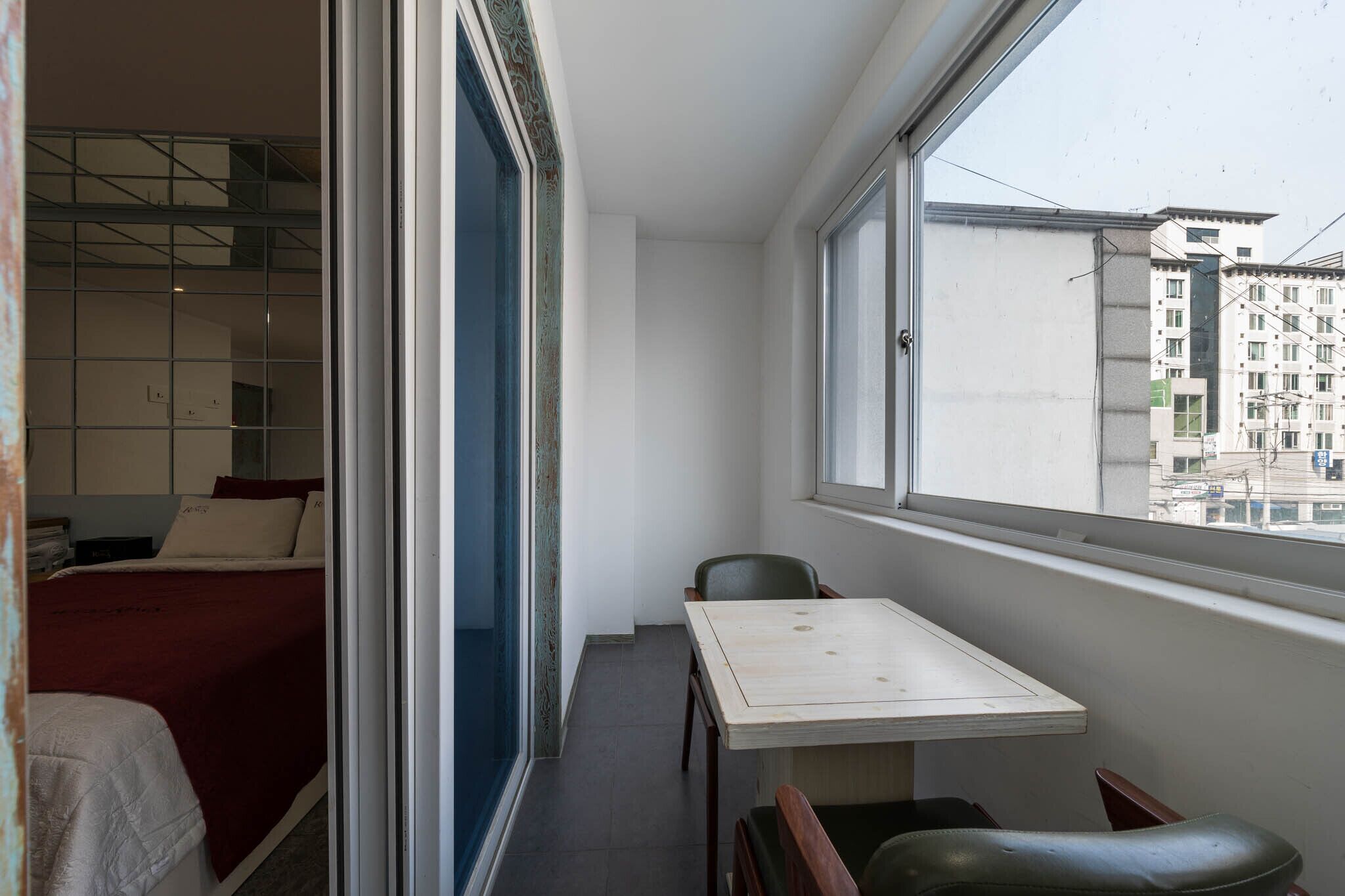 A small room with a bed, a table, and a chair. There is a window with a view of buildings outside.