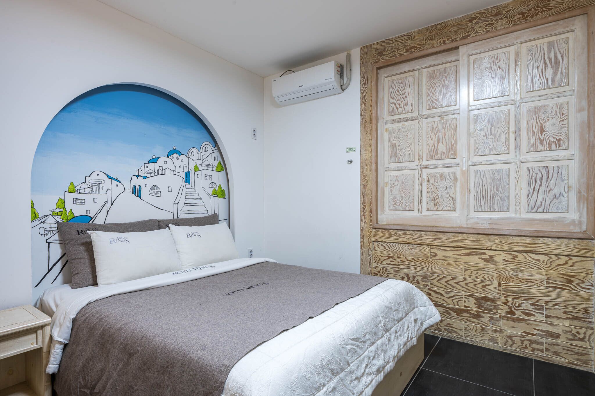 A bedroom with a bed, a wall-mounted air conditioner, a wooden panel wall, and a large circular window.