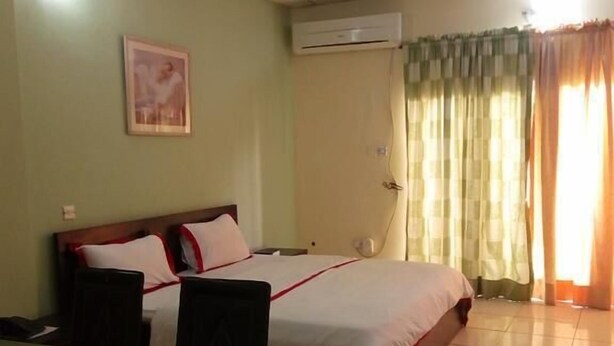 A hotel room with a bed, a desk, and a chair. There is a picture on the wall and an air conditioning unit above the bed. - The Weavers Hotel (Abuja)