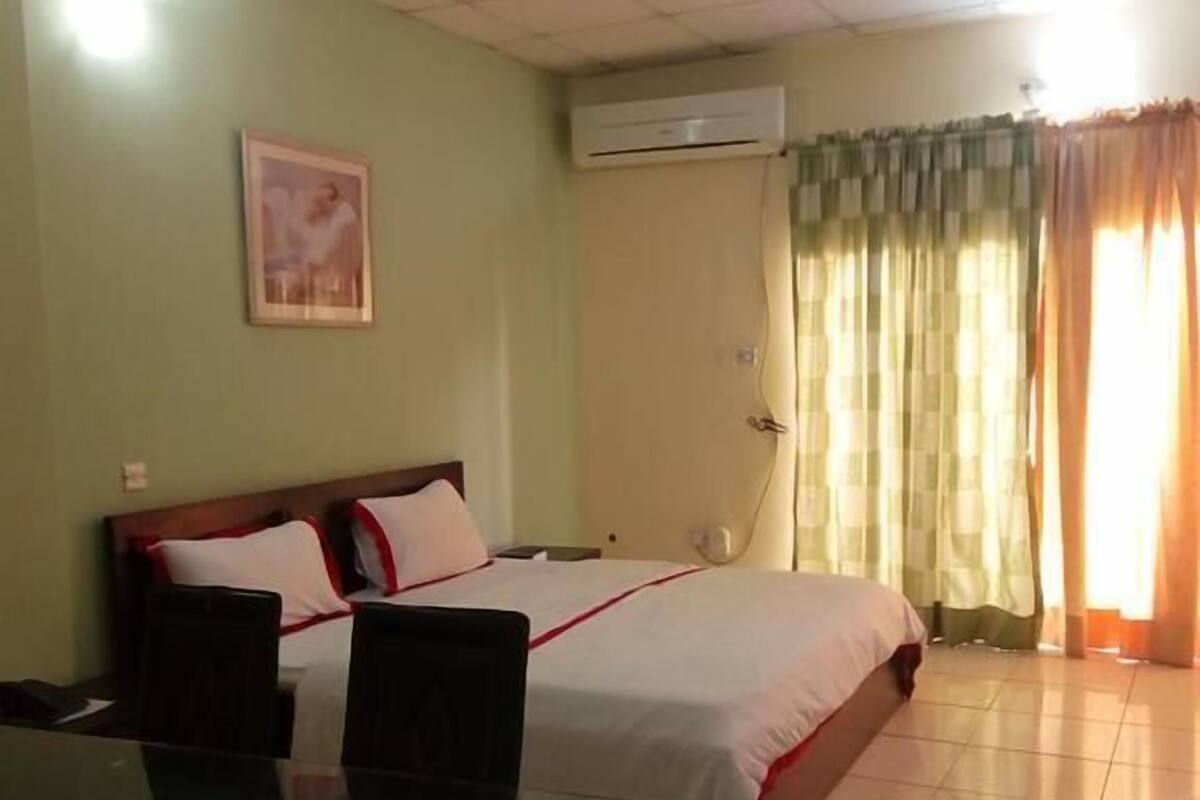 A hotel room with a bed, a desk, a chair, an air conditioning unit, and a window with curtains.