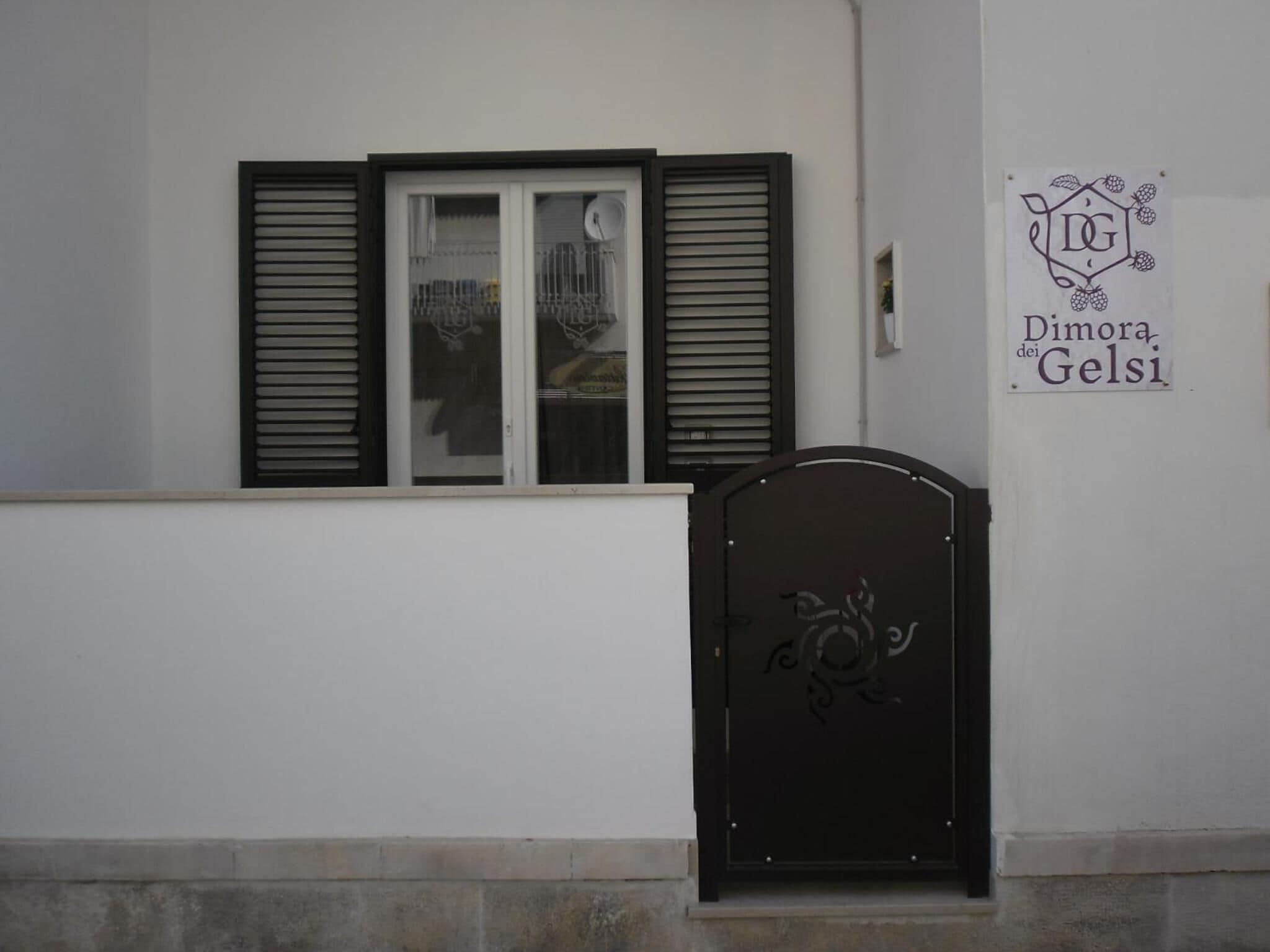 Une façade d’immeuble avec un portail noir, une fenêtre avec des volets et une enseigne indiquant « Dimora dei Gelsi ».