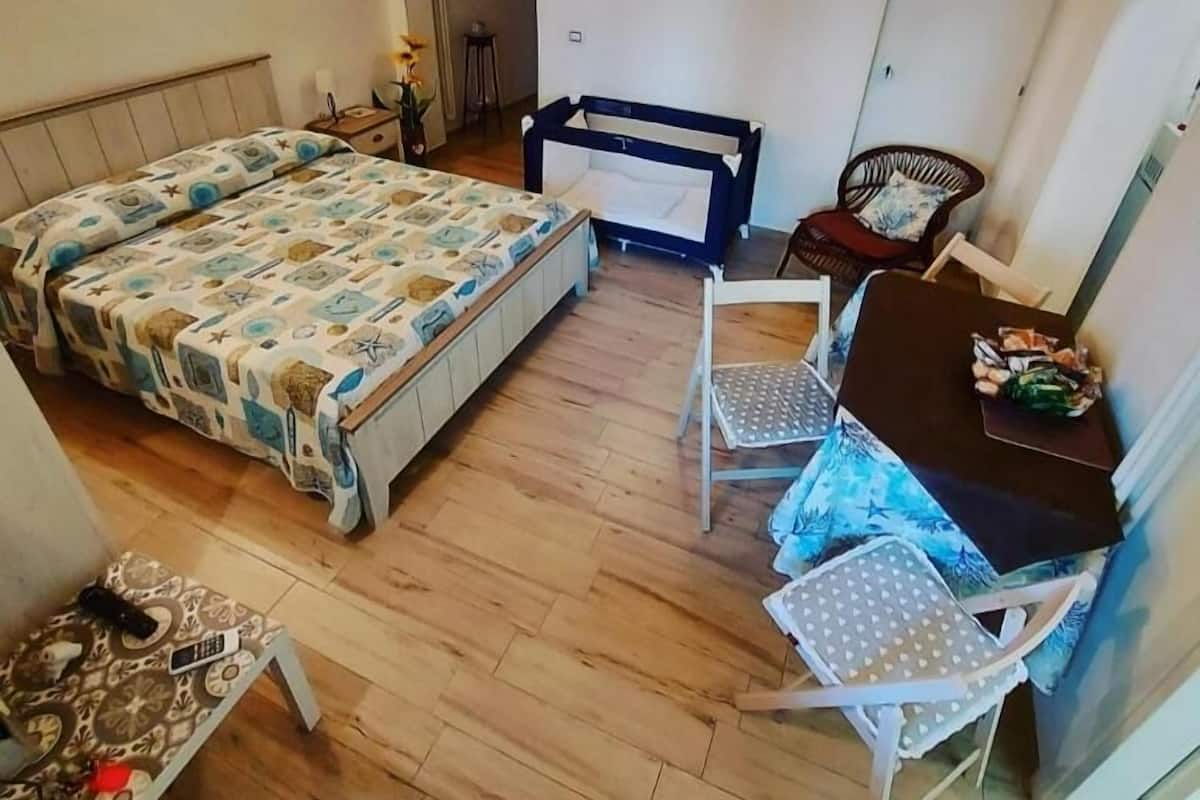 Un dormitorio con una cama, un escritorio, dos sillas, una mesita con objetos y una puerta.