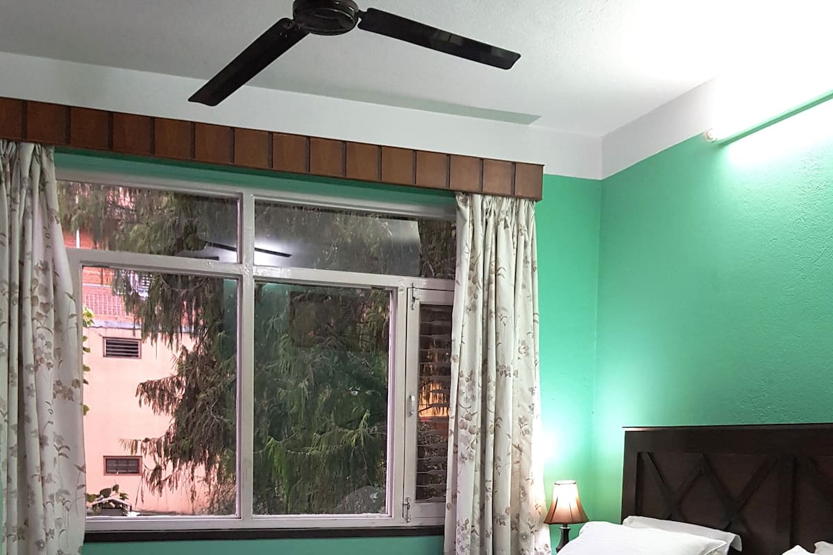 Un dormitorio con cama, ventilador de techo, ventana con cortinas y mesita de noche con lámpara.