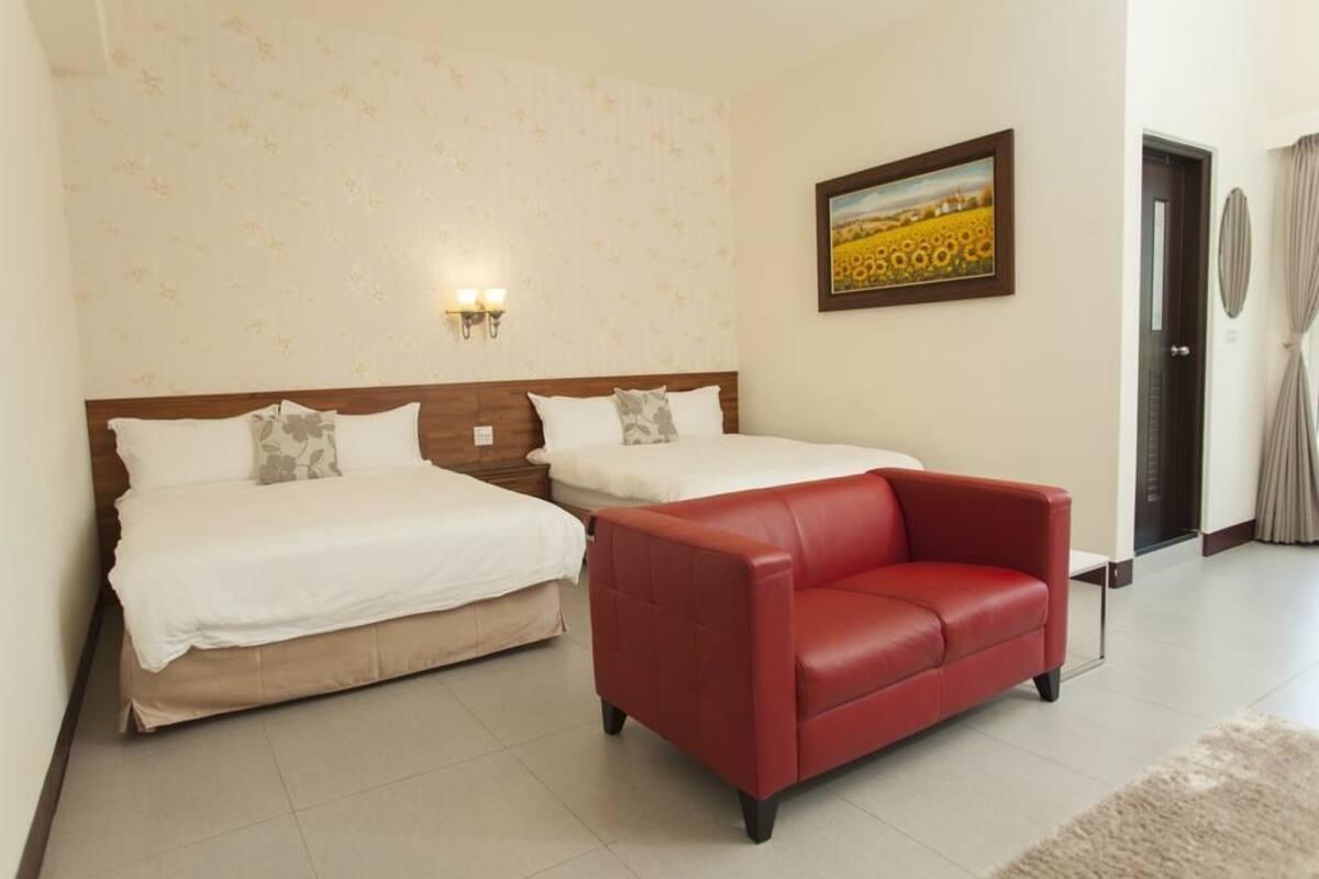 A hotel room with two beds, a red leather sofa, a painting on the wall, and a door with curtains.