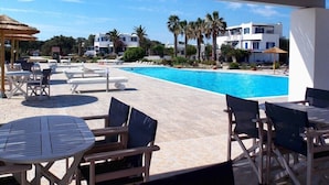 Un área junto a la piscina con sillones de descanso y mesas, una piscina azul cristalina y edificios modernos al fondo.
