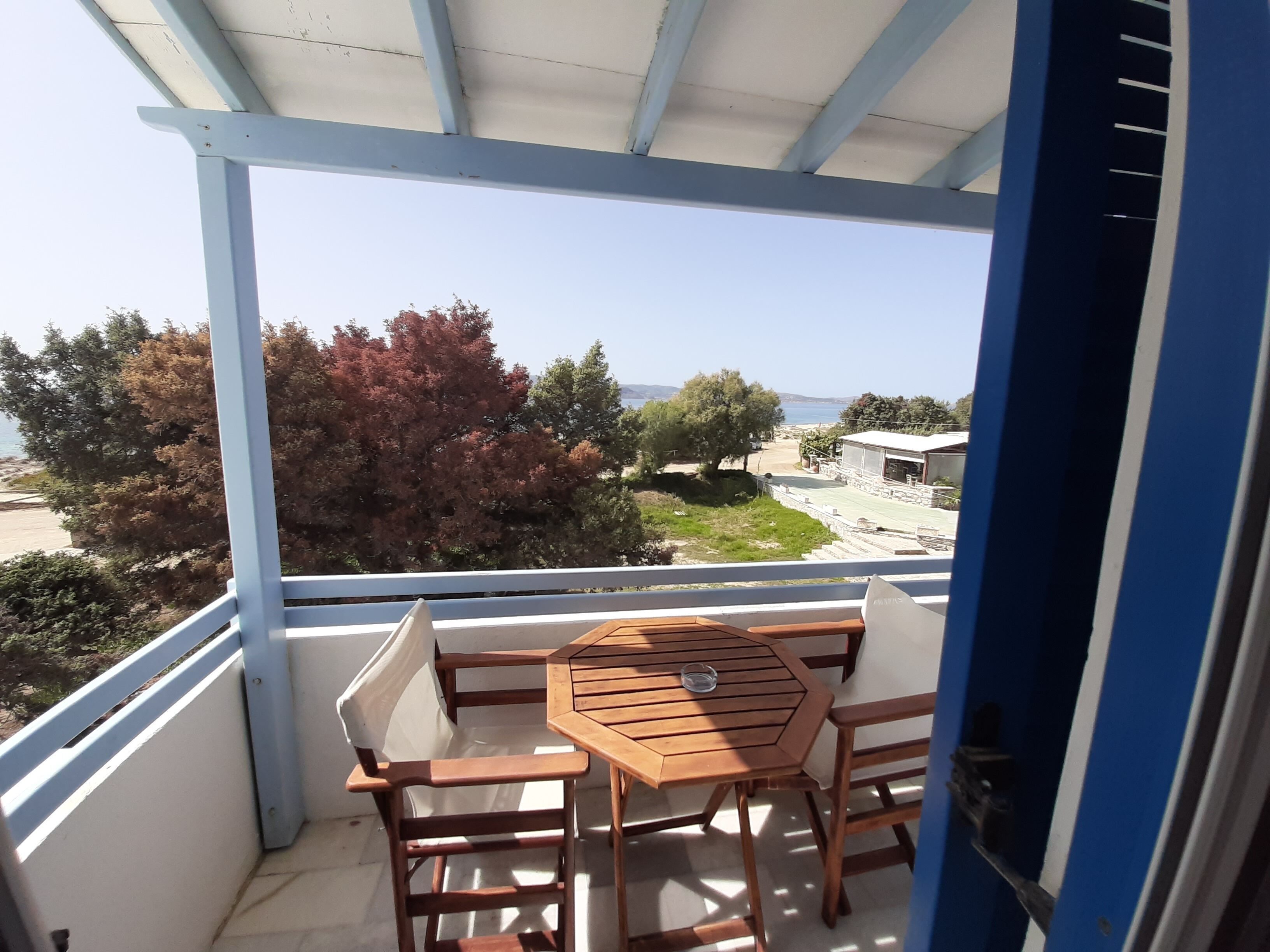 A balcony with a wooden table and chairs, overlooking a coastal area with trees and a building.