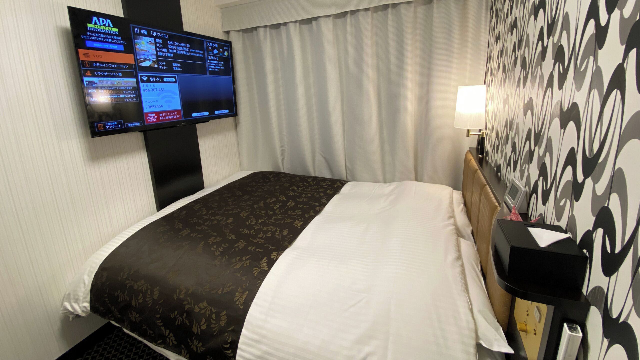 A hotel room with a bed, a TV mounted on the wall, a desk with a lamp, and a patterned wall.