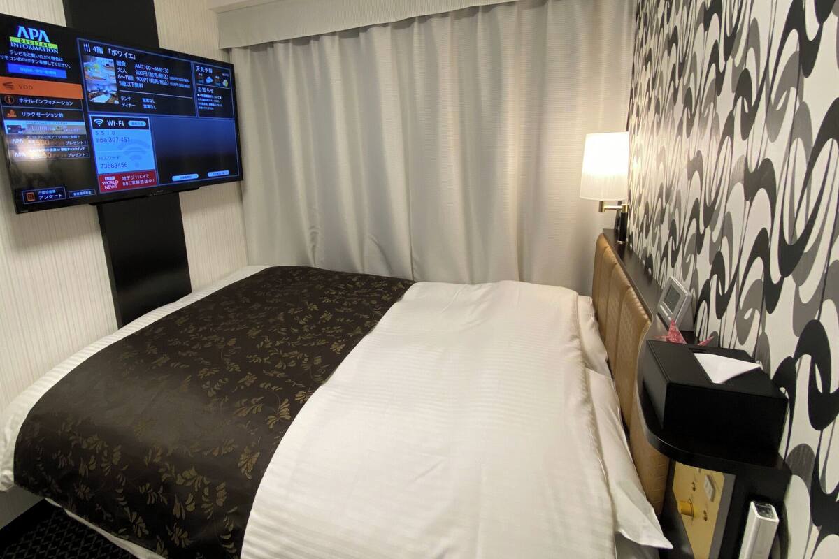 A hotel room with a bed, a TV mounted on the wall, a desk with a lamp, and a patterned wall.