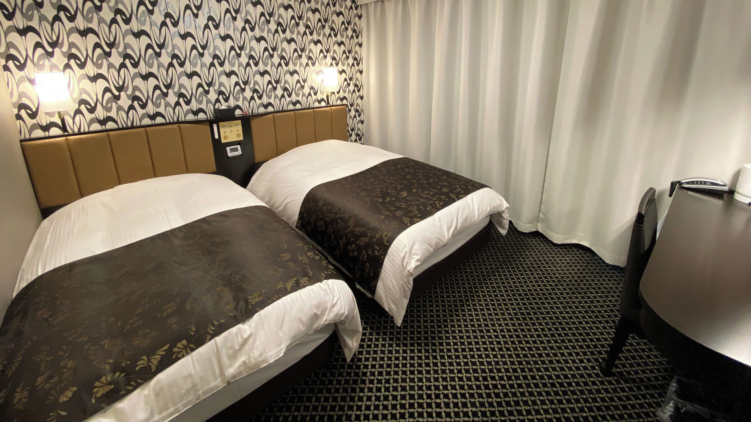 A hotel room with two beds, a headboard with controls, a table, and a patterned wall.
