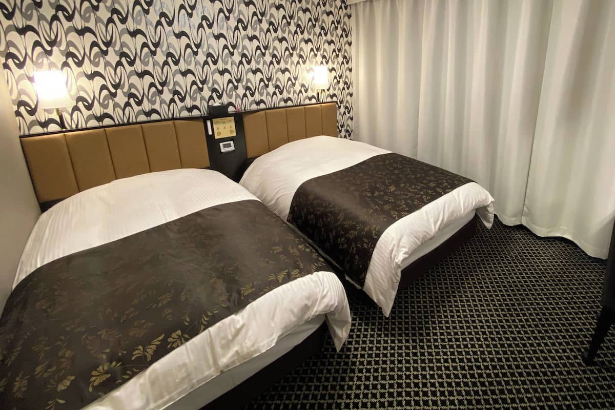 A hotel room with two beds, a headboard with controls, a table, and a patterned wall.