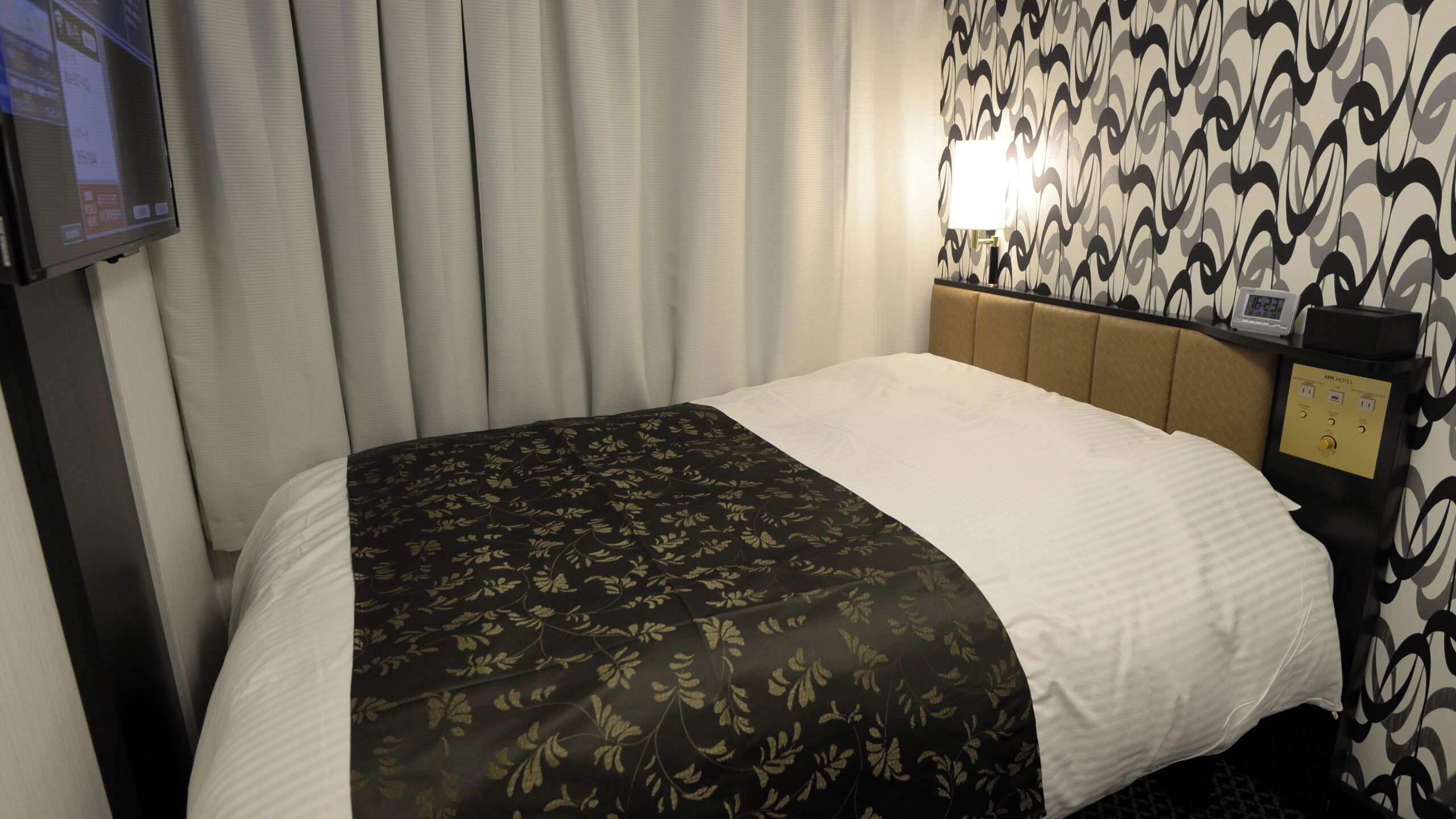 A hotel room with a bed, curtains, a TV, a wall clock, and a control panel.