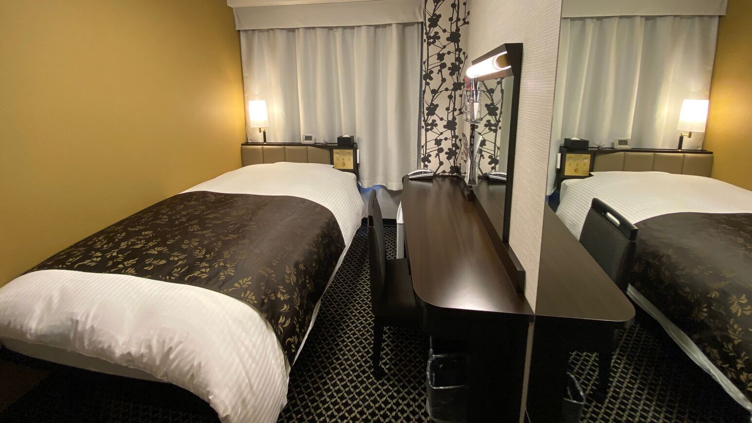Habitación de hotel con dos camas individuales, un escritorio, una silla, un espejo, una lámpara y una ventana con cortinas.
