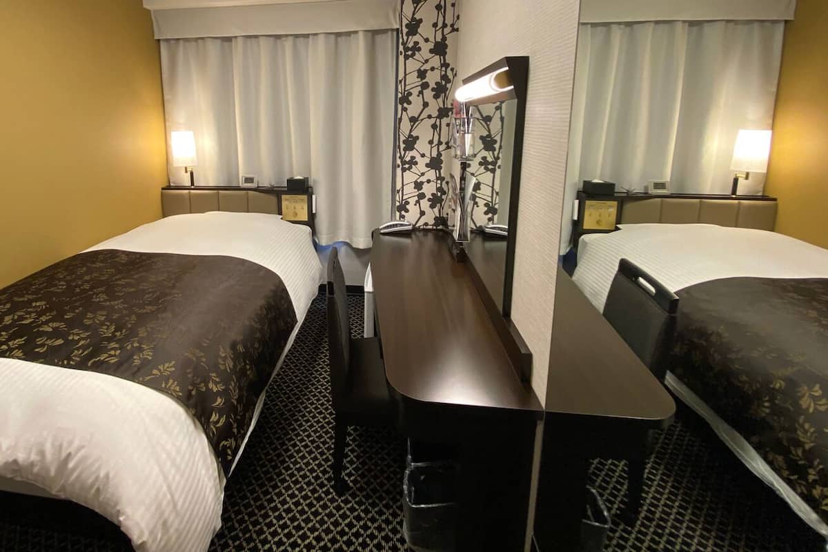 Habitación de hotel con dos camas individuales, un escritorio, una silla, un espejo, una lámpara y una ventana con cortinas.