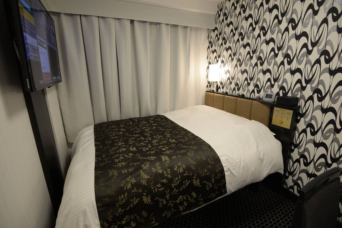 A hotel room with a bed, a television, a headboard with a control panel, and patterned wallpaper.