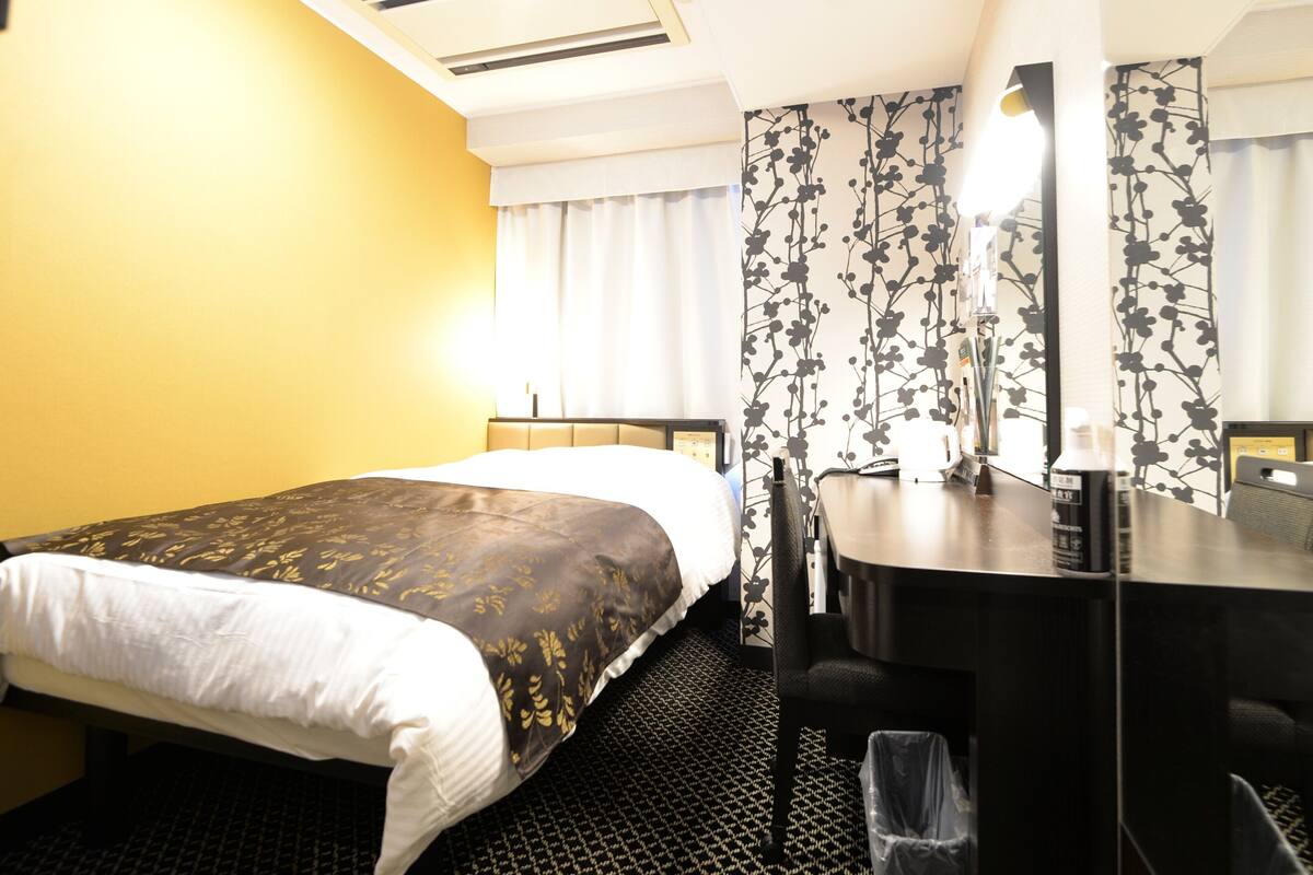 A hotel room with a bed, a desk, a chair, a trash bin, and a window with curtains.