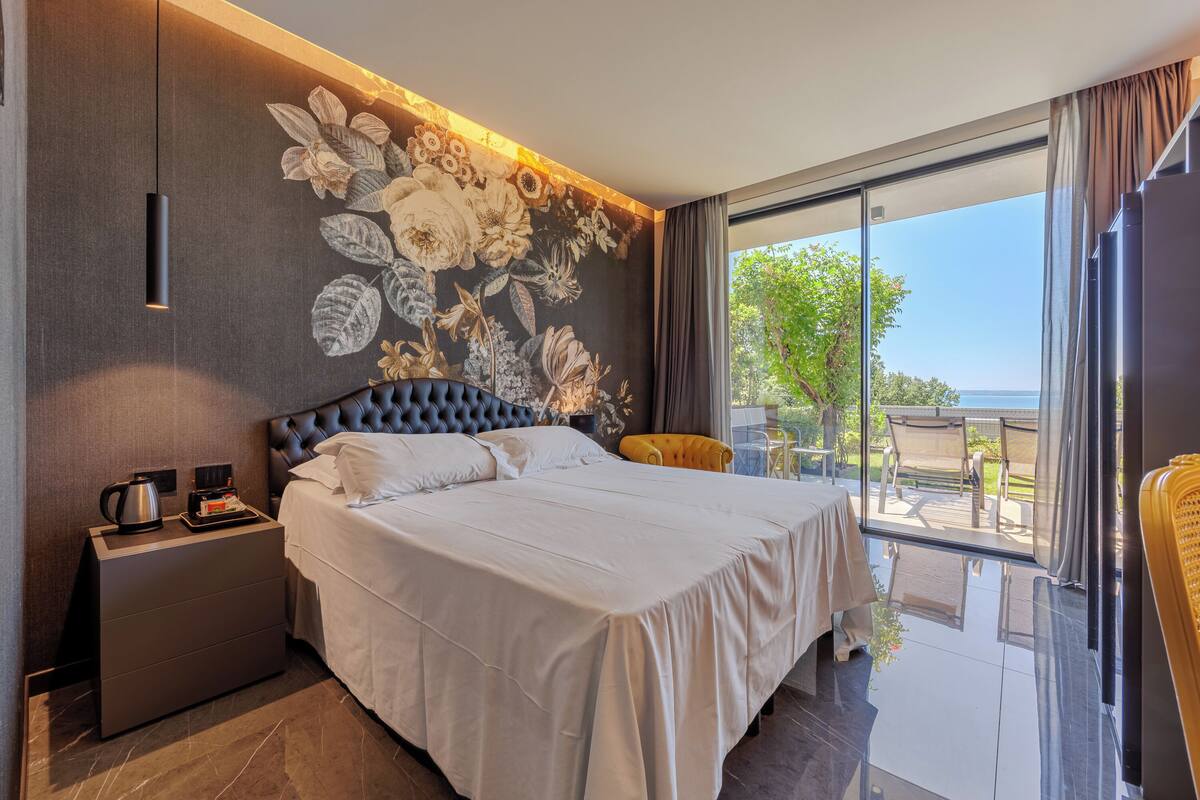 Una habitación de hotel moderna con una cama grande, una mesita de noche, una cafetera y vistas al exterior a través de un ventanal amplio.