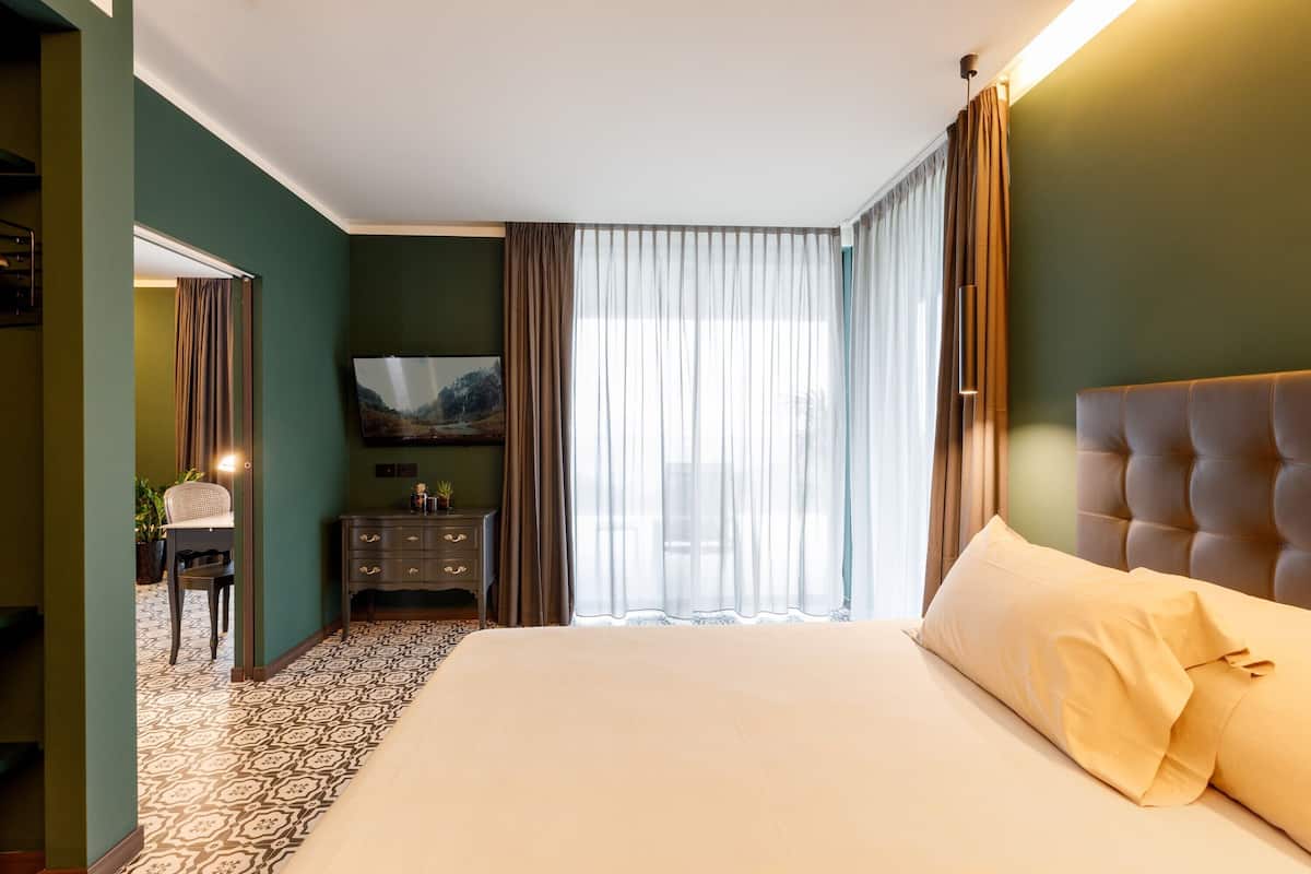 Habitación de hotel con una cama grande, un televisor en la pared, un escritorio con una silla y vistas al exterior a través de cortinas translúcidas.