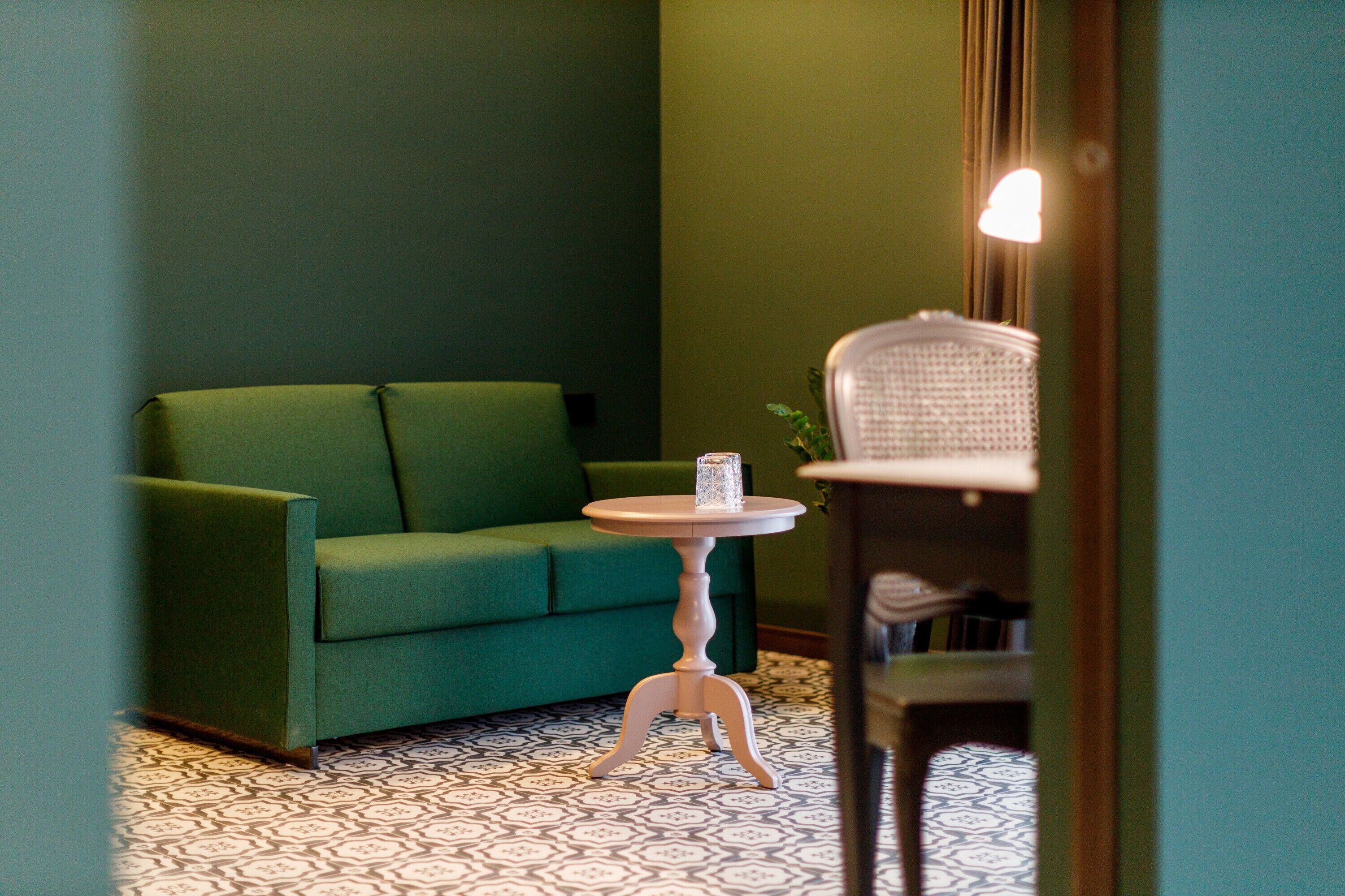 A green sofa, a small round table with a candle, and a chair in a room with patterned flooring.