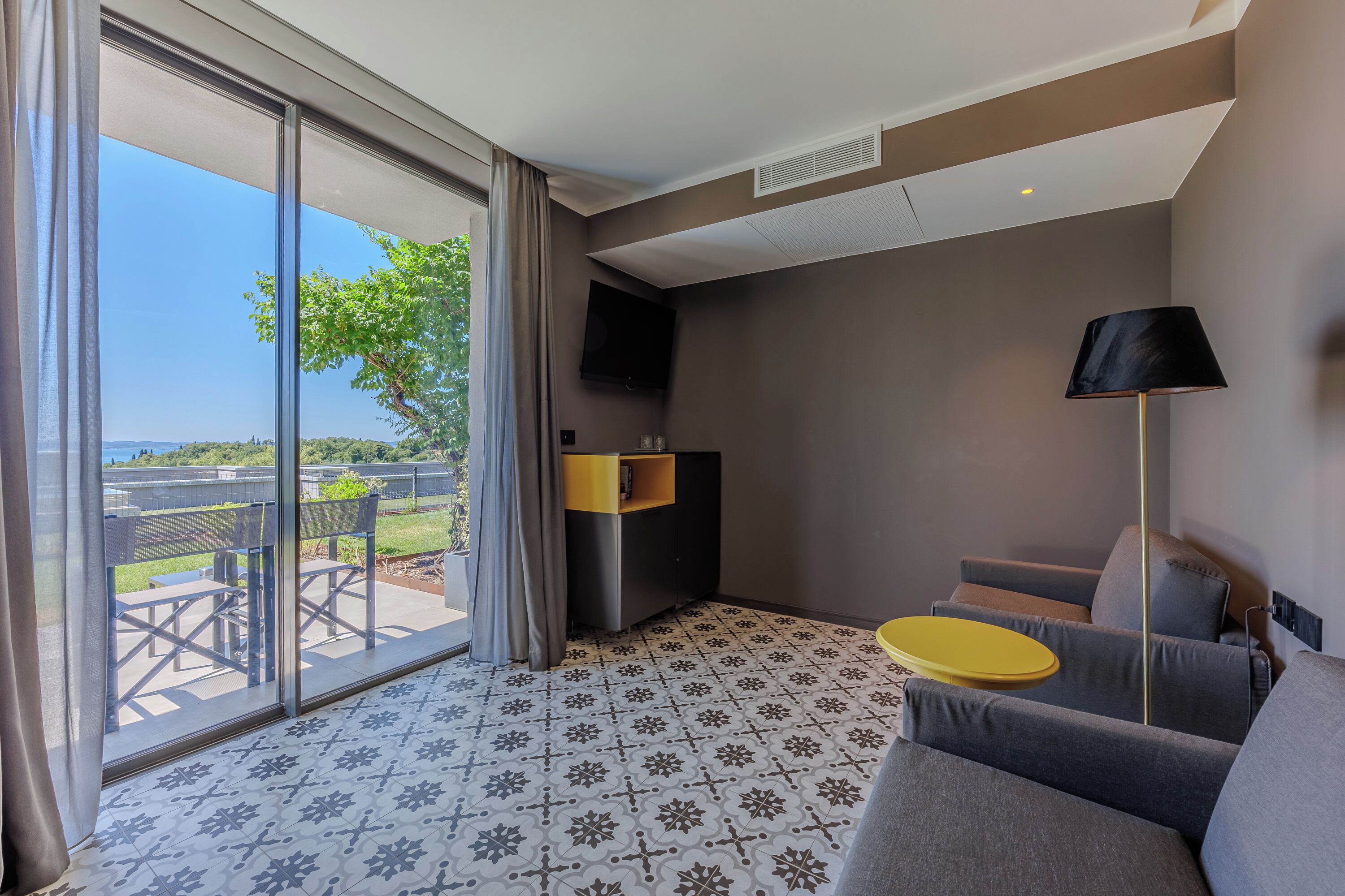 Une chambre d’hôtel moderne dotée d’une grande porte-fenêtre coulissante donnant sur un espace extérieur, d’un canapé, d’une petite table jaune et d’une lampe noire.