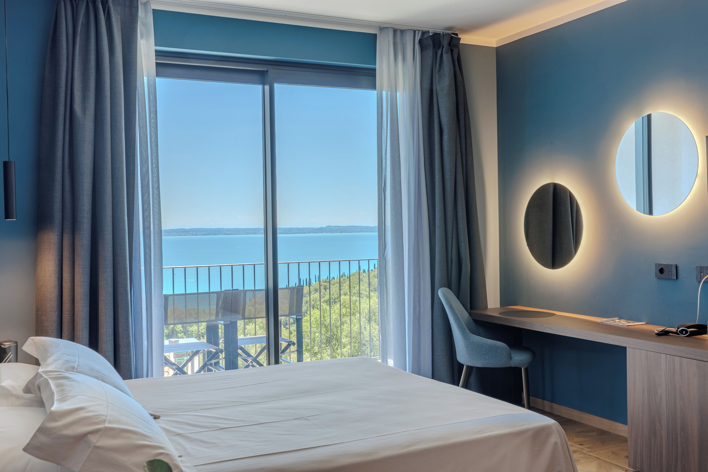 A hotel room with a large bed, a desk with a chair, a round mirror, and a view of the sea through a balcony.