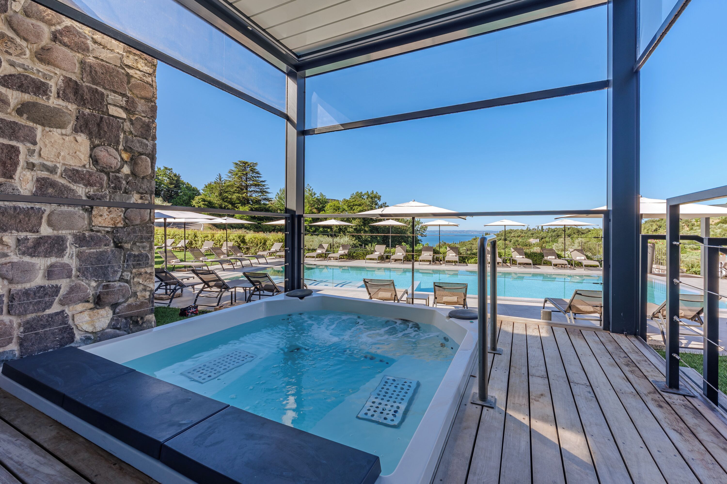 A hot tub area with a stone wall, wooden deck, and a pool with lounge chairs and umbrellas.