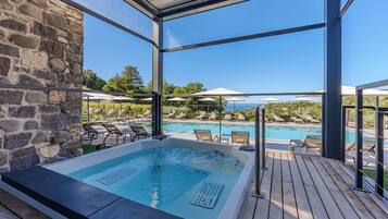 A hot tub area with a stone wall, wooden deck, and a pool with lounge chairs and umbrellas.