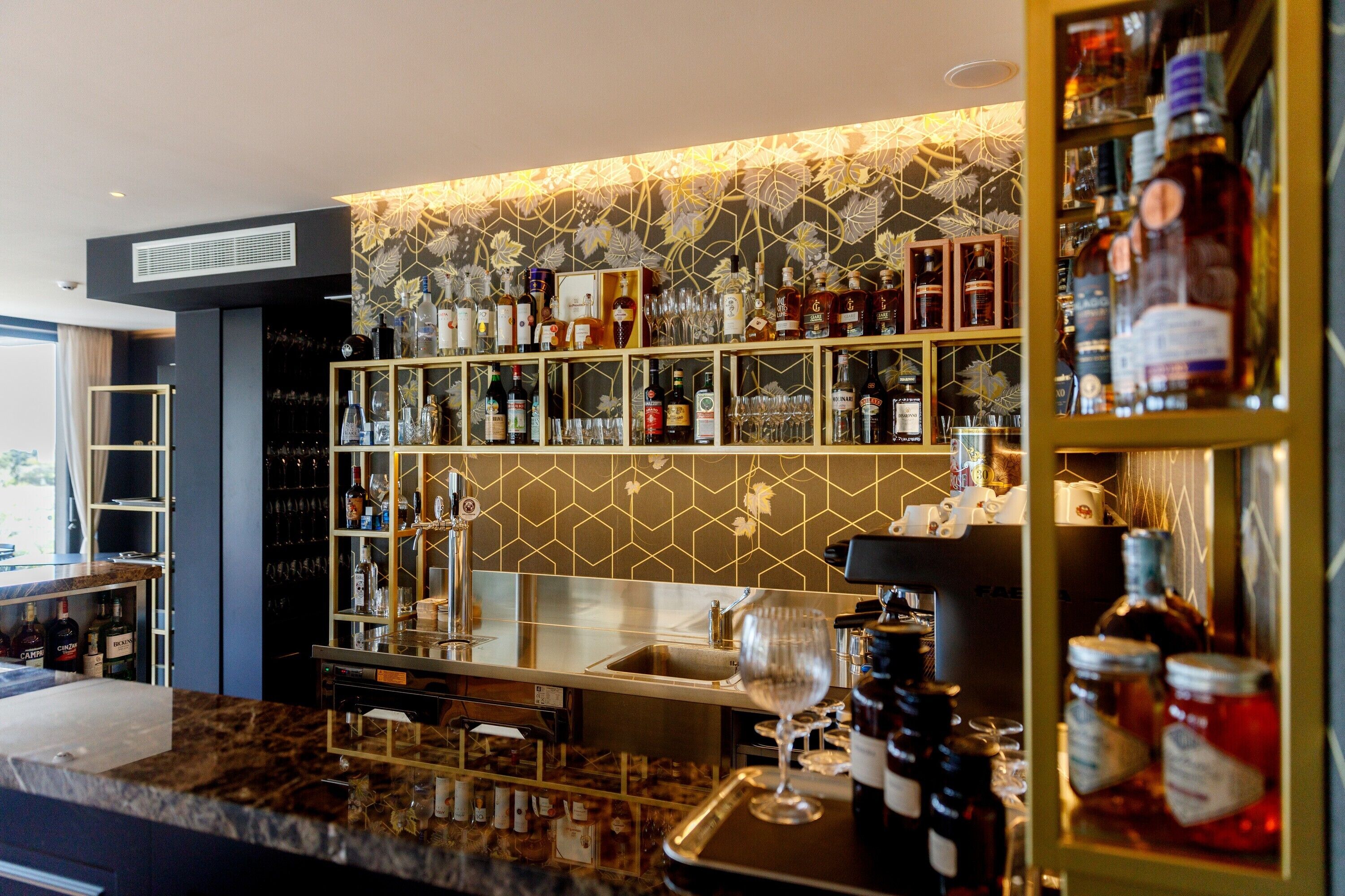 Zona de bar con estantes llenos de botellas de licores, vasos y una copa de vino sobre la barra.
