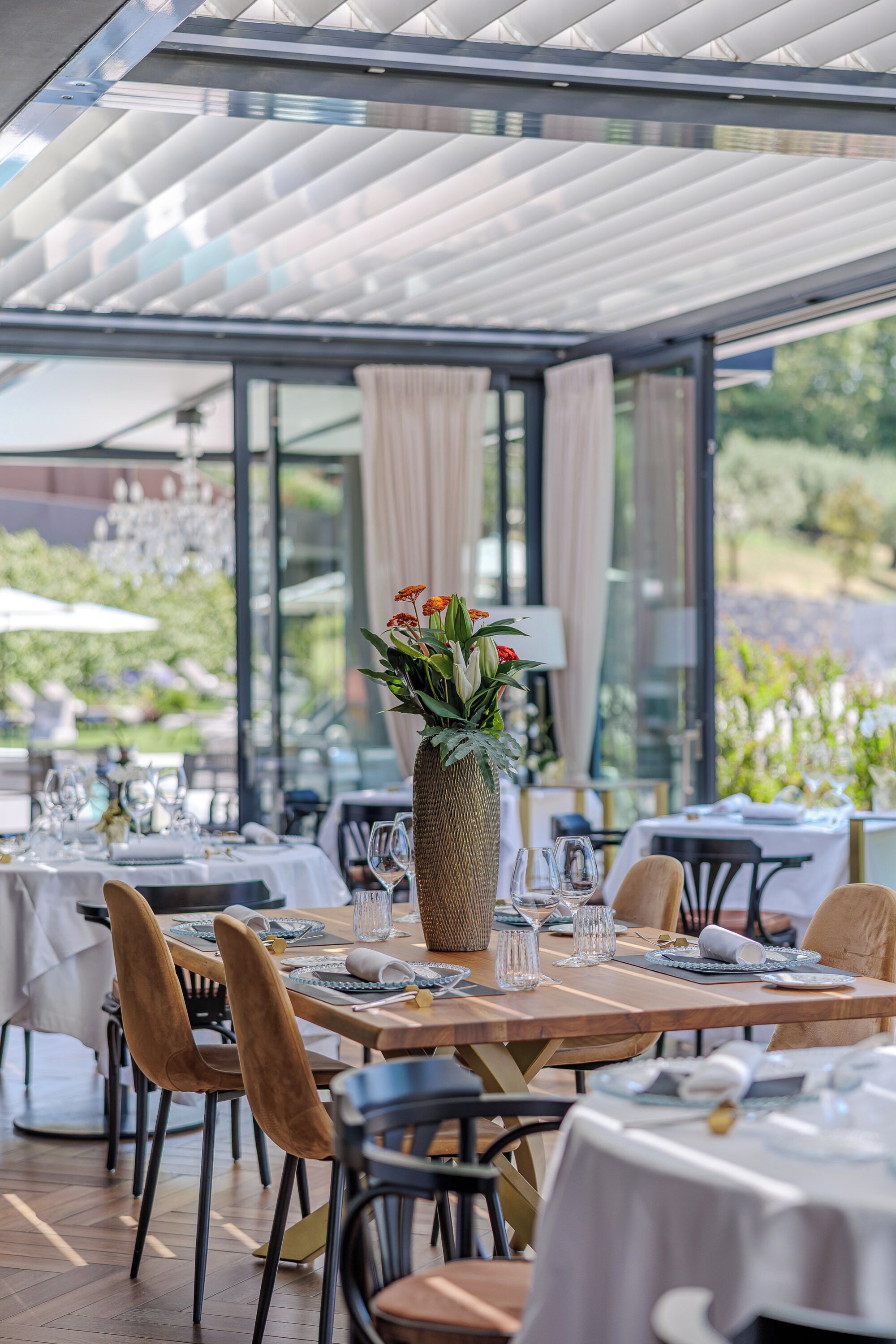 Un área de comedor al aire libre con mesas dispuestas para dos, destacando un jarrón grande con flores, sillas de madera y un techo de cristal.