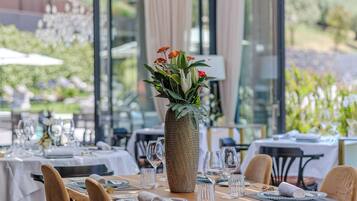 Un área de comedor al aire libre con mesas dispuestas para dos, destacando un jarrón grande con flores, sillas de madera y un techo de cristal.