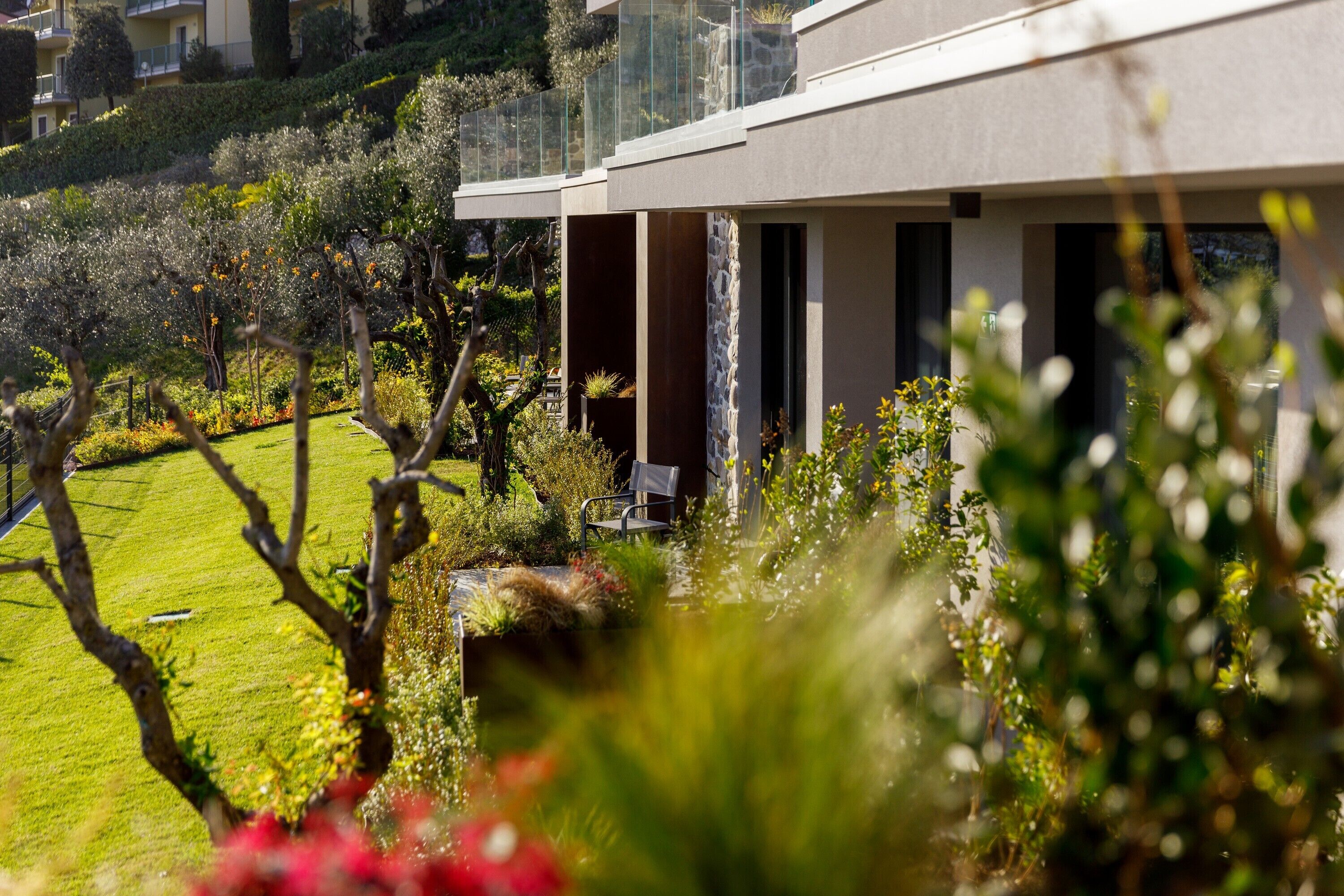 A modern building with a glass balcony overlooking a well-maintained garden with various plants and a lawn.