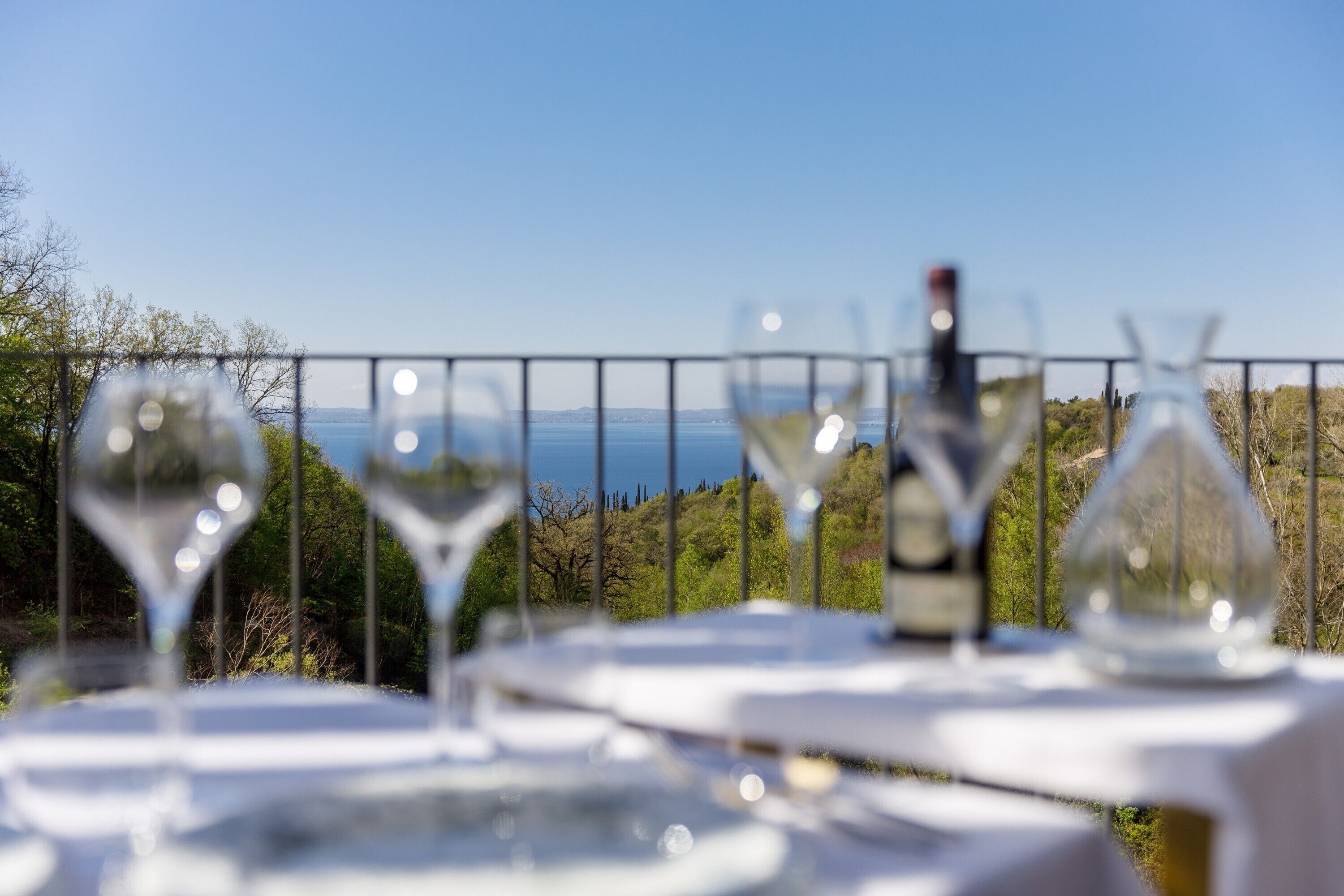 Una mesa con copas de vino y una botella, frente a una vista panorámica de un cuerpo de agua y colinas.