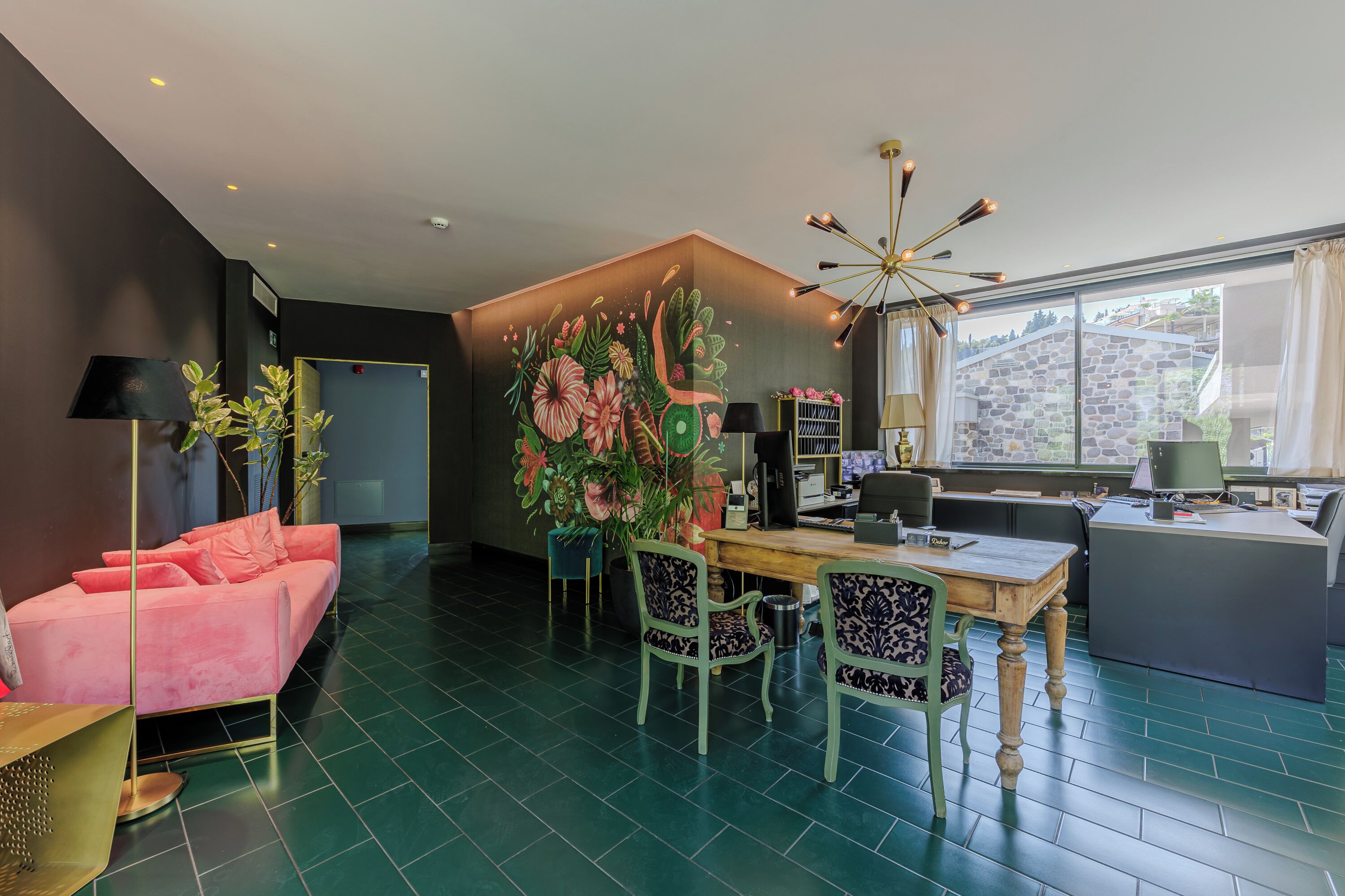 Una sala de estar moderna con un sofá rosa, una mesa de comedor con sillas, un mural floral grande y vistas a un edificio de piedra a través de amplios ventanales.