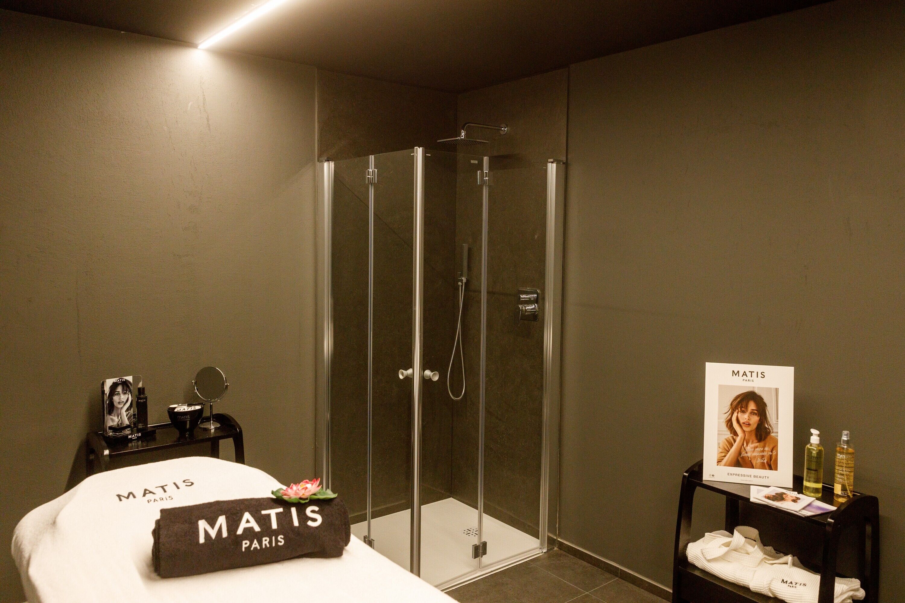 Een moderne badkamer met een douchecabine van glas, een bed met een handdoek met de tekst 'Matis Paris' en een klein tafeltje met toiletartikelen.