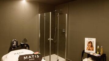 Et moderne badeværelse med en brusekabine i glas, en seng med et håndklæde mærket "Matis Paris" og et lille bord med toiletartikler.
