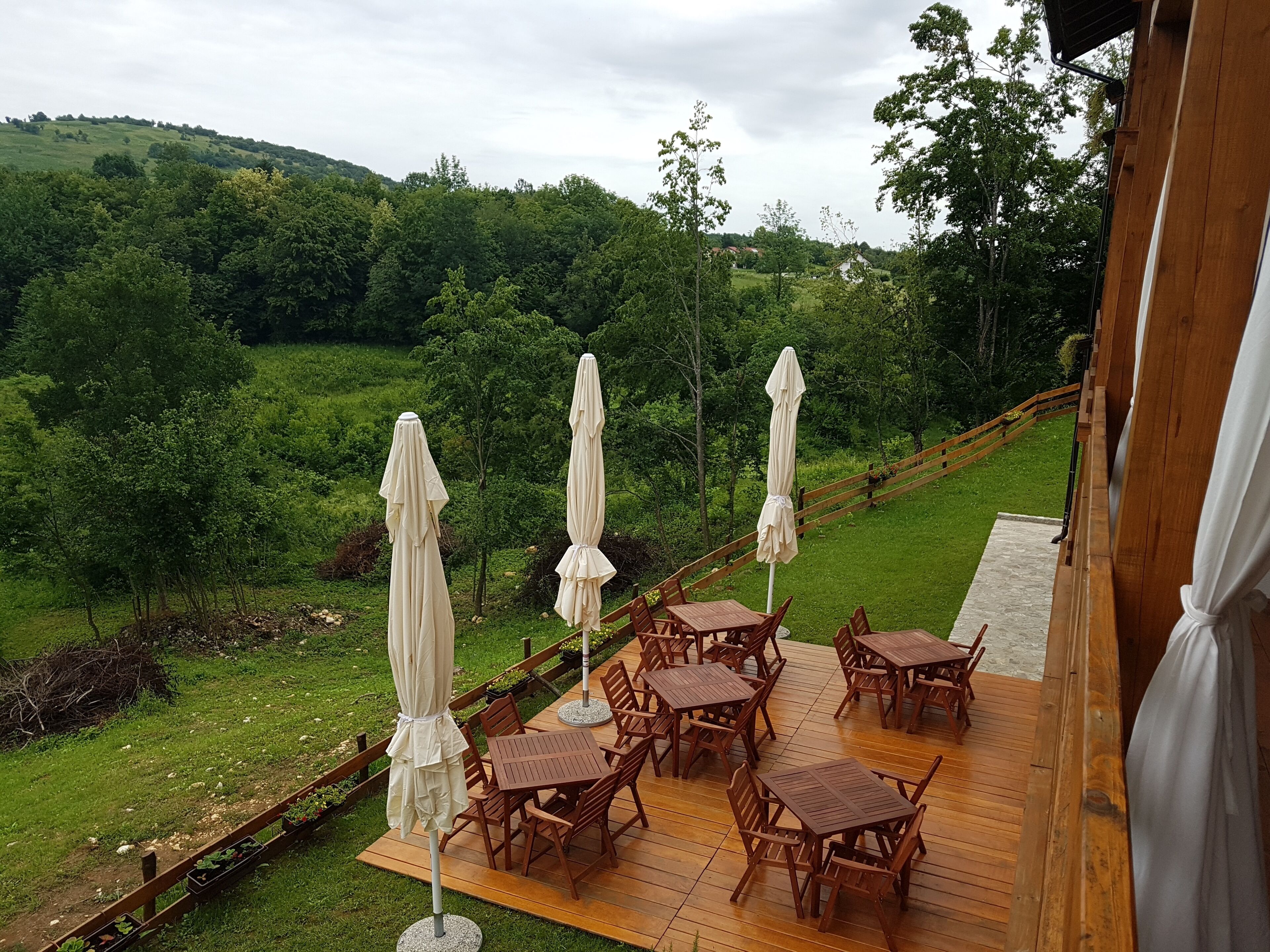 A wooden deck with outdoor tables and chairs, surrounded by lush greenery and a view of rolling hills.