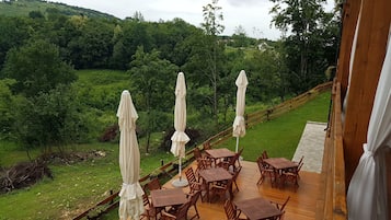 A wooden deck with outdoor tables and chairs, surrounded by lush greenery and a view of rolling hills.