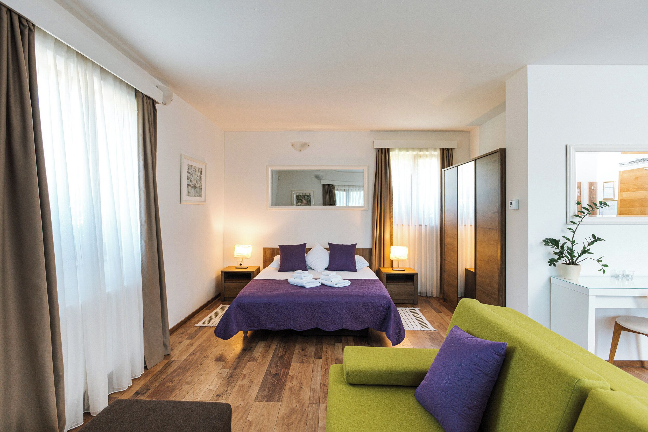 A hotel room with a large bed, two bedside tables with lamps, a desk, a green sofa, and a potted plant.