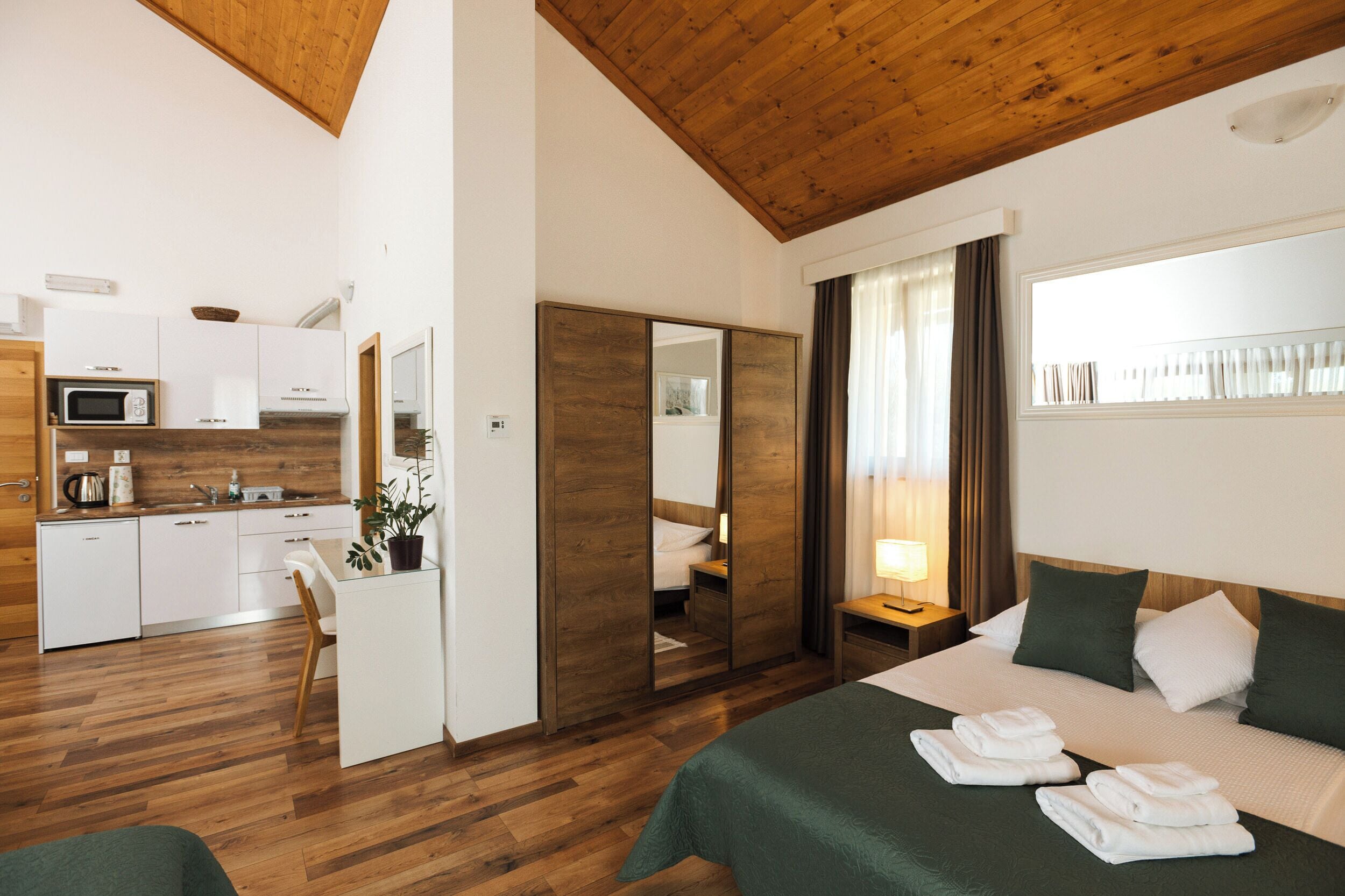A hotel room with a bed, a bedside table, a mirror, a small kitchen area with a microwave, and a wooden ceiling.