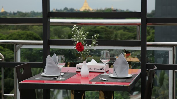 Mesa para dos con un jarrón de flores y copas de vino, con vistas a un paisaje urbano que incluye una pagoda dorada.