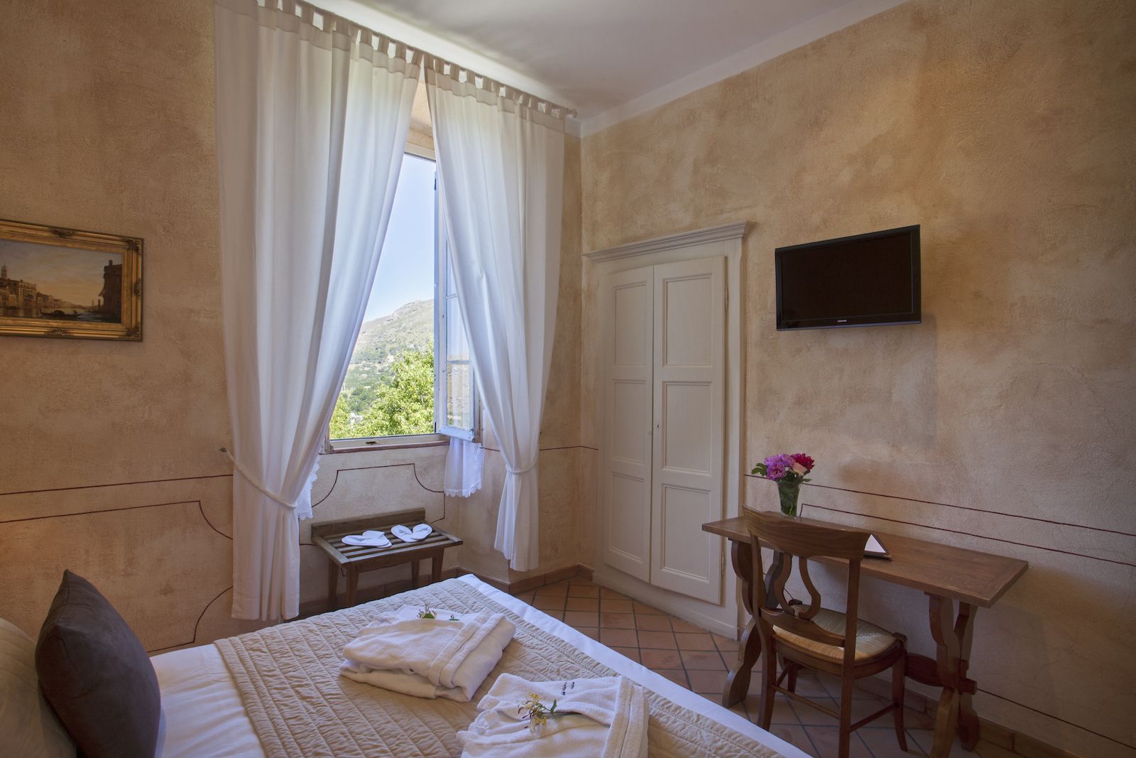 A bedroom with a bed, a bedside table with towels, a chair, a desk with a vase of flowers, a painting on the wall, and a TV mounted on the wall.