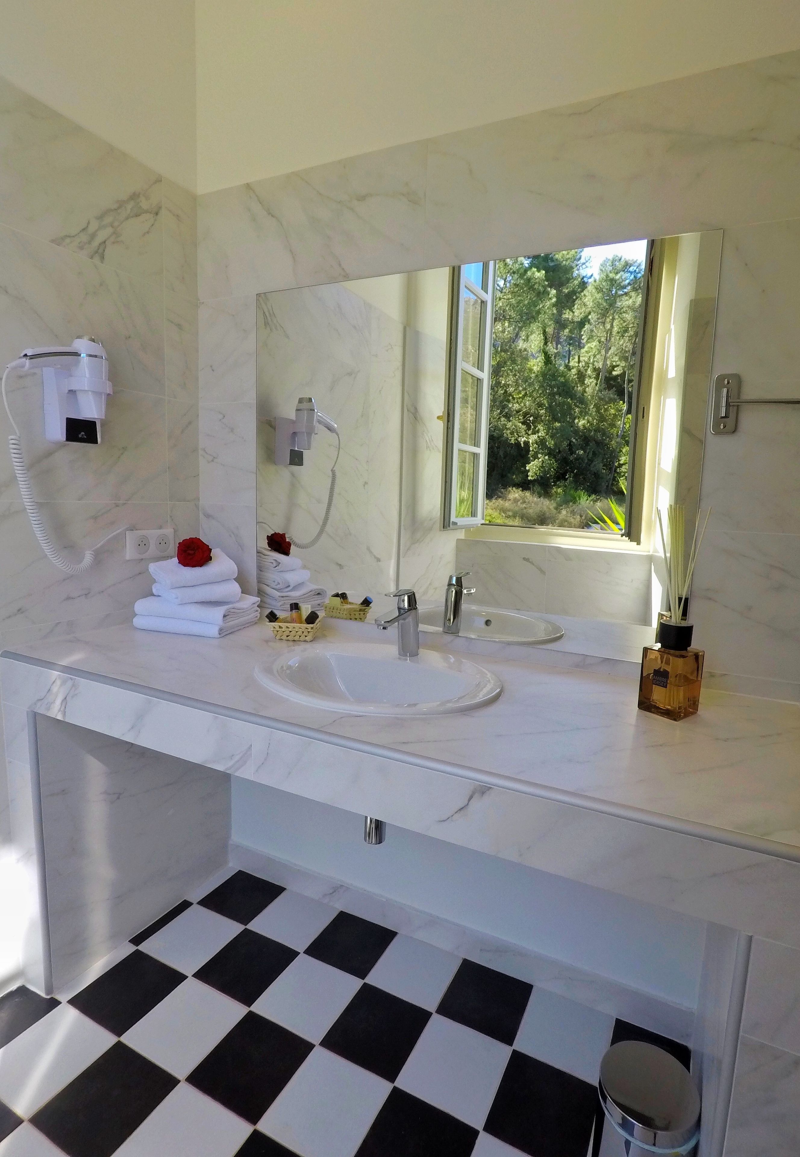 A bathroom with a white marble countertop, two sinks, a large mirror, a hairdryer, and a window with a view of greenery.