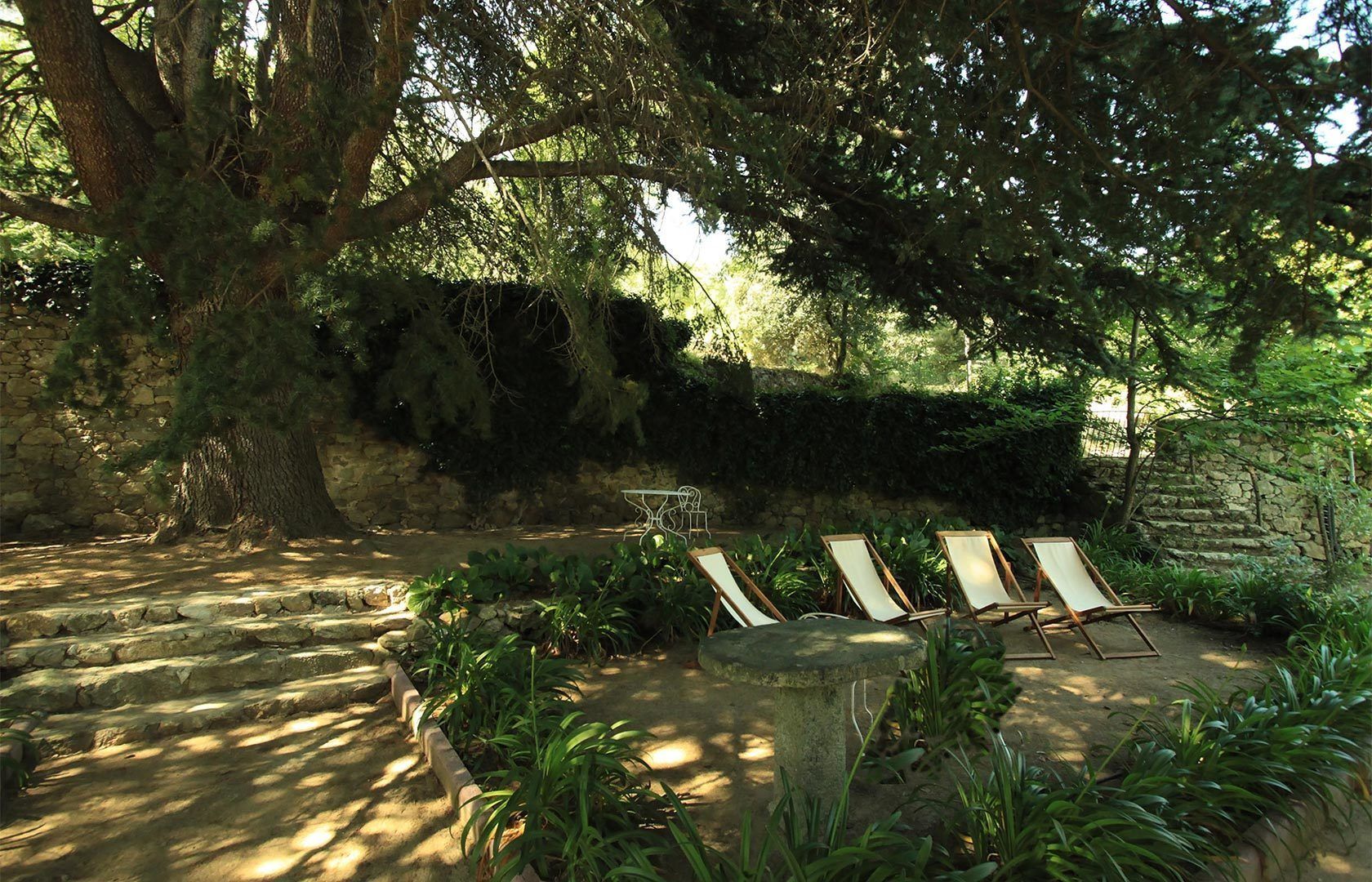 A stone pathway leading to a shaded seating area with lounge chairs and a table, surrounded by lush greenery and a stone wall.