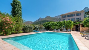 Una piscina rectangular con agua azul cristalina, rodeada por una solana con baldosas. Hay sillas de descanso y un edificio con balcón al fondo.