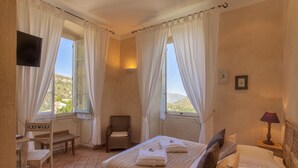 Un dormitorio con cama, una silla, un escritorio, televisor y ventana con cortinas.