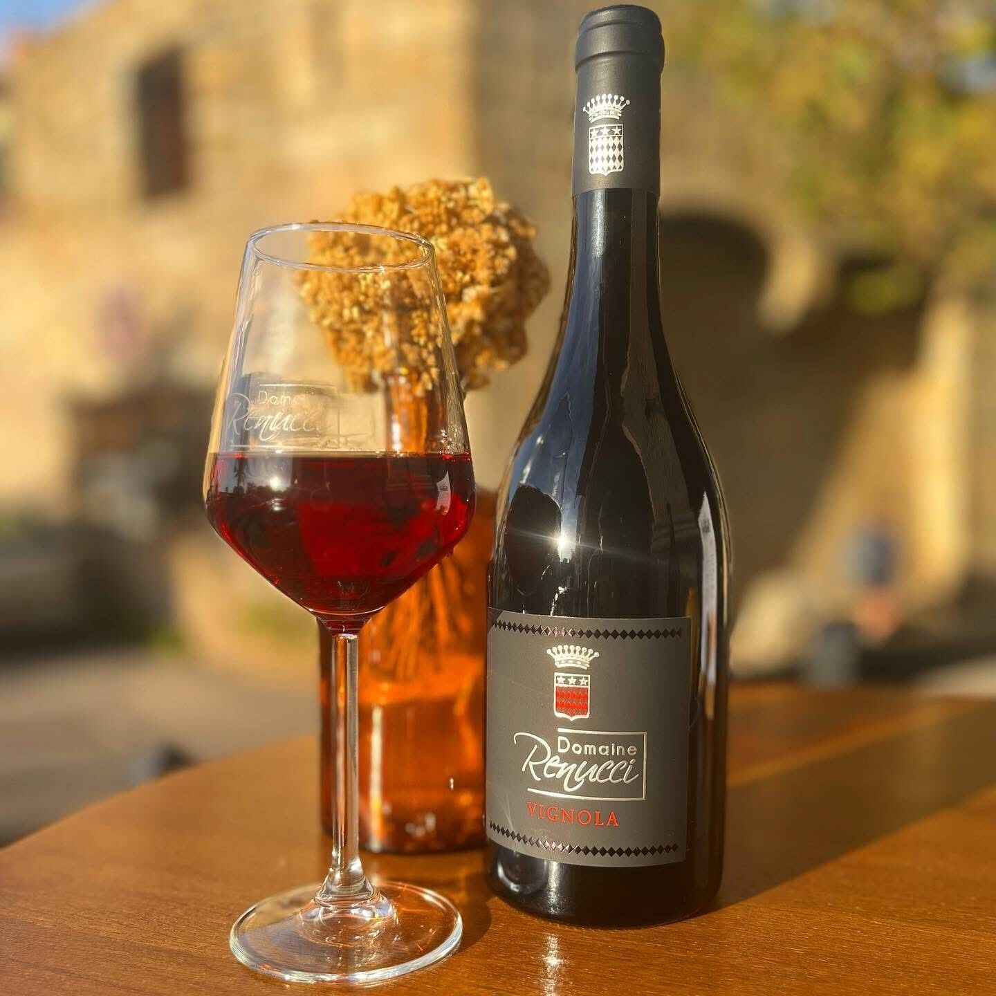 A bottle of Domaine Renucci Vignola wine and a glass of red wine on a wooden table with a blurred background.