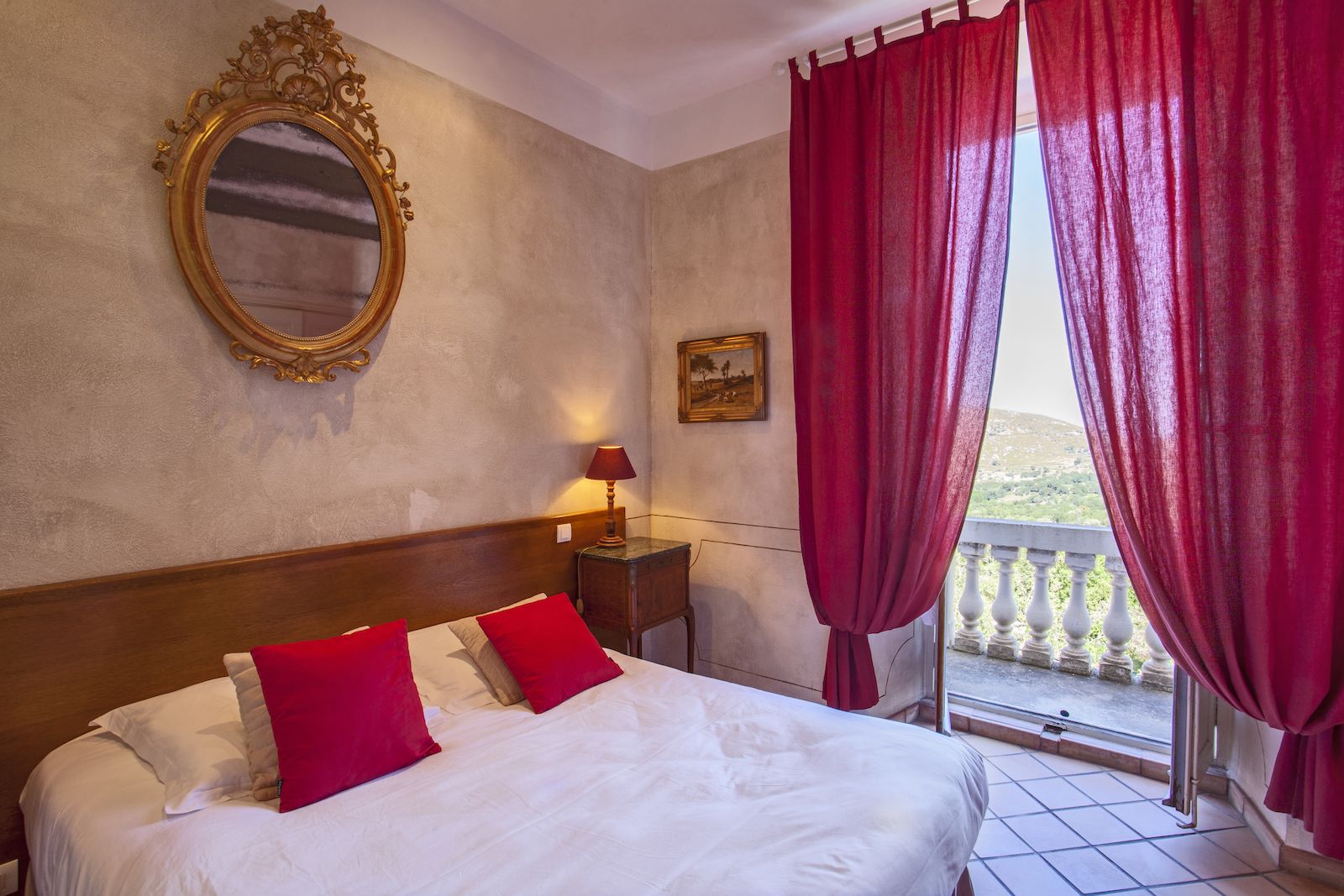 A hotel room with a large bed, red pillows, a wooden headboard, a nightstand with a lamp, a framed picture on the wall, and a balcony with red curtains.