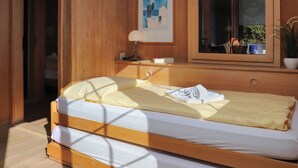 A wooden-framed bed with a white mattress and a yellow blanket, a bedside table with a lamp, a framed picture on the wall, and a window with a view of greenery outside. - Jungfrau a d Ledi by Interhome (Wengen)
