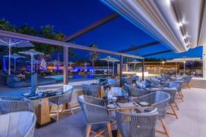 An outdoor dining area with tables and chairs, umbrellas, and a pool in the background. - Imperial Palace Hotel (Rethymno)