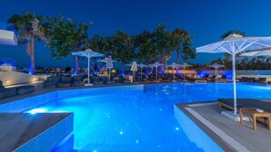 A swimming pool with lounge chairs and umbrellas, surrounded by palm trees and night lighting.