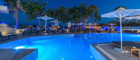 Una piscina con sillas de descanso y sombrillas, rodeada de palmeras e iluminación nocturna.